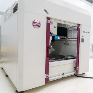 Optotech MCP 1501 polisher in IACTEC's south cleanroom A large white machine with two big doors (polishing machine for large surfaces) in a white room with high ceilings (IACTEC's south clean room).