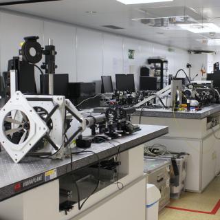 Vista general de una de las salas (A) del laboratorio de óptica. Laboratorio alargado con mesas metálicas con agujeros y diversos elementos ópticos y mecánicos encima de ellas
