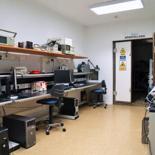 Vista general del laboratorio de compatibilidad electromagnética con bancos de trabajo y ordenadores y vista al fondo la sala de la puerta de entrada a la sala apantallada