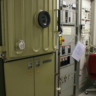 Vista de la evaporadora y de su electrónica asociada en el laboratorio Vista de la evaporadora y de su electrónica asociada en el laboratorio. Máquina con una puerta reforzada y un ojo de buey, y dos grandes racks con componentes electrónicos, botones e indicadores para el control de la máquina.